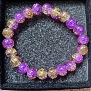 Puede incluir: Un brazalete de cuentas con piedras preciosas de color púrpura y amarillo alternadas. Las cuentas redondas y translúcidas se presentan en una caja negra. Las piedras presentan variaciones naturales de color. Cada cuenta mide aproximadamente 0,8 cm de diámetro.