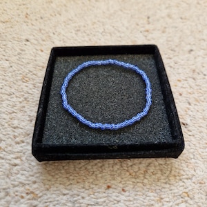 May include: A circular bracelet composed of small, round, light blue beads with thin, white, diagonal striped patterns. The bracelet is displayed resting flat inside a square, black, foam-lined jewelry box with a textured surface.