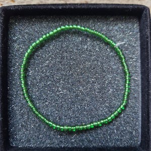Könnte beinhalten: Ein zartes Armband aus kleinen, leuchtend grünen Perlen. Das Armband ist kreisförmig und liegt in einer schwarzen, samtgefütterten Schmuckschatulle, was einen auffälligen Kontrast erzeugt. Die Perlen scheinen gleichmäßig angeordnet zu sein, was auf ein handgefertigtes oder sorgfältig gearbeitetes Design hindeutet.
