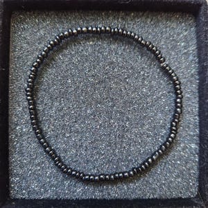 May include: A black beaded bracelet presented in a black velvet jewellery box. The bracelet is composed of small, round, dark beads strung together. The beads have a subtle sheen, reflecting light. The jewellery box has a textured, grey interior.