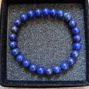 May include: A bracelet composed of spherical, deep blue beads. The beads are strung together, forming a circular shape. The bracelet is presented in a black, velvet-lined box, providing a strong contrast with the vivid blue beads.