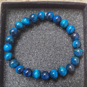May include: A blue beaded bracelet against a dark backdrop. The bracelet features polished, spherical beads in diverse blue hues, strung together to form a circle. The bracelet is presented in a black box.
