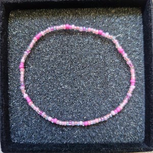 May include: A delicate beaded bracelet featuring a mix of pink and purple beads. The bracelet is displayed on a black velvet surface, showcasing its colors and design. This piece is a stylish accessory.