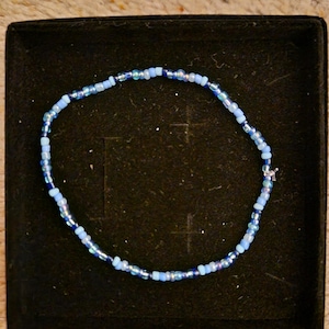 Puede incluir: Un brazalete de cuentas azules y azul claro con un cierre plateado. El brazalete se muestra en una caja de terciopelo negro.