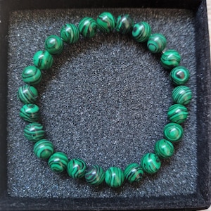 May include: A beaded bracelet featuring round, green malachite stones with dark green and black banding. The bracelet is displayed in a black, velvet-lined box, showcasing the natural patterns of the gemstones.