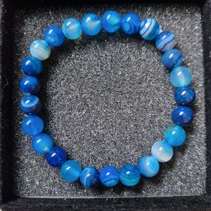 Peut inclure: Un bracelet de perles bleues avec des nuances de bleu variées. Les perles rondes sont enfilées ensemble, créant une forme circulaire. Le bracelet est présenté dans une boîte noire avec un intérieur texturé. Les perles mesurent environ 0,8 cm de diamètre.