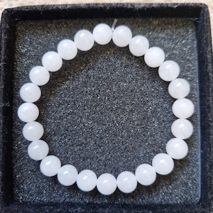 May include: A white beaded bracelet composed of round, opaque beads. The bracelet is displayed in a black square box with a textured interior. The beads are strung together to form a circular shape, suitable for wrist wear.