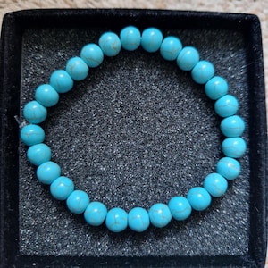 May include: A turquoise beaded bracelet presented in a black velvet jewellery box. The bracelet features round, light blue beads with delicate darker veining. The beads are strung together, forming a circular design.