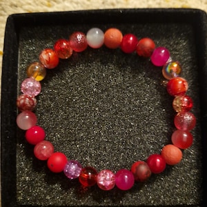 May include: A bracelet made of red, pink, and clear beads of varying sizes and textures. Some beads are matte, others glossy. The bracelet is presented in a black box, highlighting the range of colours. The beads are likely between 0.5 and 1 cm in diameter.