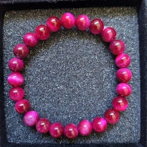 Może przedstawiać: Żywa różowa bransoletka z koralików. Bransoletka składa się z okrągłych, wypolerowanych koralików w różnych odcieniach różu i magenty. Koraliki są nawleczone razem, tworząc pełne koło. Bransoletka jest prezentowana na ciemnoszarej powierzchni w czarnej tacy.
