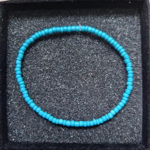 May include: A turquoise beaded bracelet in a black box. The bracelet is made of small, round beads, strung together to form a circle. The beads are a vibrant shade of blue, presented in a jewellery box.
