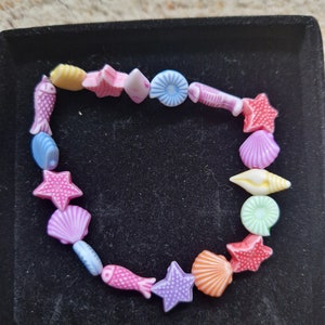 May include: A colorful beaded bracelet featuring various sea-themed charms. The bracelet includes charms shaped like fish, starfish, and seashells in shades of pink, blue, yellow, and orange. The bracelet is displayed on a black surface.