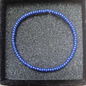 May include: A circular bracelet composed of small, matte, royal blue beads arranged in a continuous loop. The beads are uniform in size and shape, creating a simple, singular strand design that rests within a square, black, foam-lined display box.