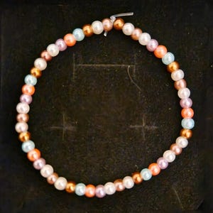 May include: A colourful beaded bracelet with a mix of beads. The bracelet includes white, peach, light blue, and gold-coloured beads. The bracelet is displayed on a black surface.