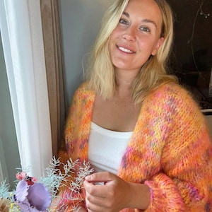 Op de afbeelding: Een vrouw draagt een kleurrijke, pluche cardigan met een wit tanktop en blauwe jeans. De cardigan heeft een mix van roze, oranje en gele tinten.