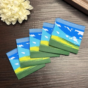 以下が含まれることがあります： 風景のデザインが施された正方形の布地のパッチが5つ。パッチには、白い雲のある青い空、黄色の畑、緑色の丘が描かれています。パッチは扇形に配置されています。