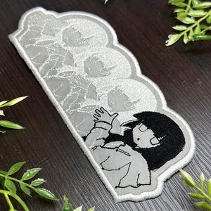 May include: A gray and white embroidered patch featuring a cartoon character with black hair and a white outfit. The patch has a cloud-like shape with a floral pattern on the top half and a character on the bottom half.