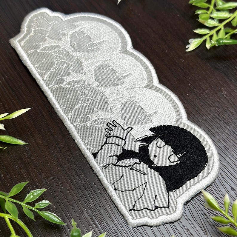 May include: A gray and white embroidered patch featuring a cartoon character with black hair and a white outfit. The patch has a cloud-like shape with a floral pattern on the top half and a character on the bottom half.