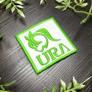 Puede incluir: Parche cuadrado blanco con borde verde y logotipo verde. El logotipo es un diseño abstracto con las letras "URA" en verde. El parche es probablemente un emblema o parche bordado para ropa o accesorios.