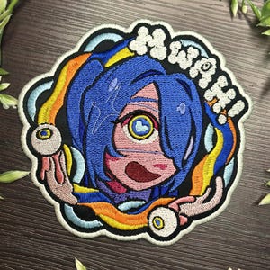 May include: Embroidered patch depicting a cartoon character with blue hair and a large eye. The character is encircled by a rainbow design and the word "Mwah!" in white. The patch has a black border.