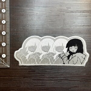 May include: Embroidered patch featuring a cartoon character with black hair and glasses, with three faded images of the same character. The patch is white with black and gray details, and is approximately 3 inches long.