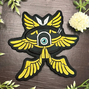 May include: A black and yellow embroidered patch with a large eye in the centre and wings on either side. The eye is blue and white.