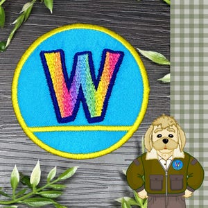 May include: A circular embroidered patch featuring a bright blue background with a large, rainbow-colored letter W in the center, outlined in dark blue, and a yellow border. Beside it is a cartoon dog wearing a brown jacket with a matching W patch.