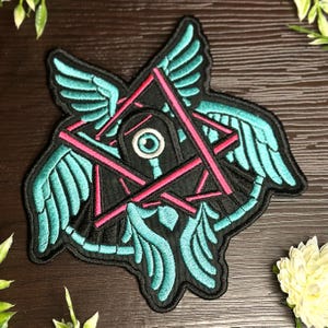 May include: An embroidered patch with a black background, featuring a pink and red star with a blue eye in the center. The star is surrounded by four turquoise wings.