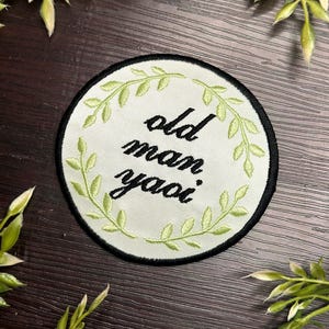 Puede incluir: Un parche circular blanco bordado con un borde de hojas verdes. El texto "old man yaoi" está bordado en negro en el parche.