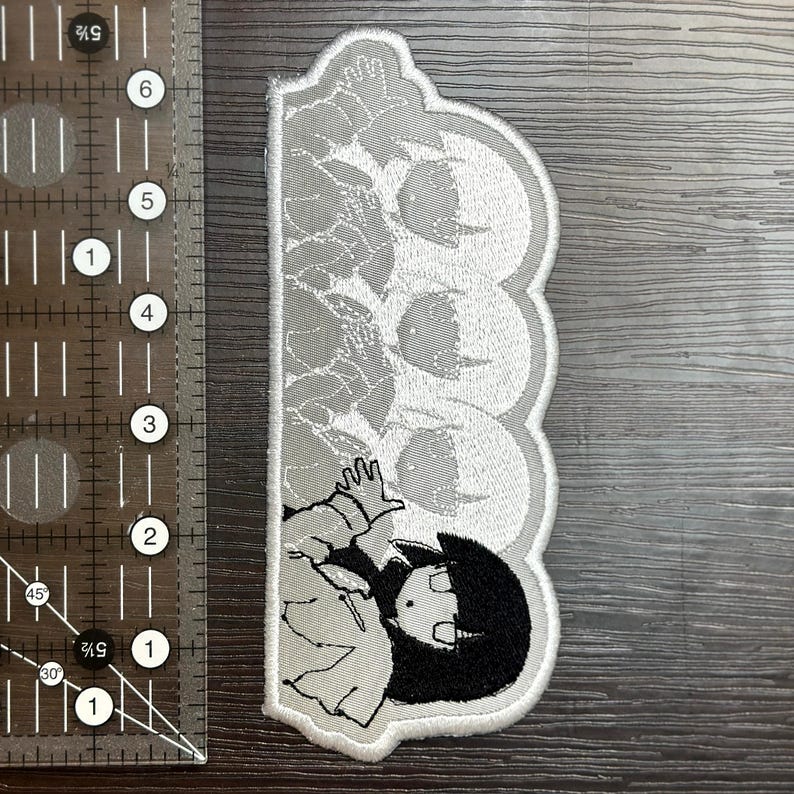May include: A fabric patch featuring a stylized anime character with black hair and a white outfit. The patch has a scalloped edge and a gray and white color scheme. The patch is approximately 5 inches in length.