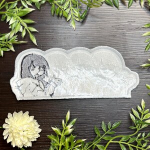 May include: A white, cloud-shaped patch with a black and white illustration of a person. The patch has a textured, velvety appearance. The illustration is of a person with long hair and large eyes. The patch is surrounded by green foliage.