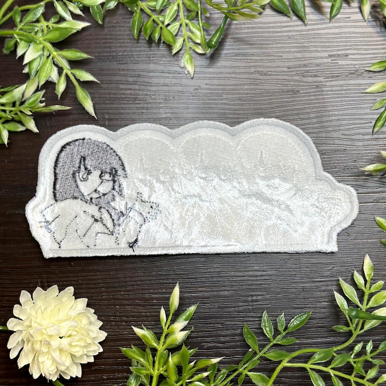 May include: A white, cloud-shaped patch with a black and white illustration of a person. The patch has a textured, velvety appearance. The illustration is of a person with long hair and large eyes. The patch is surrounded by green foliage.