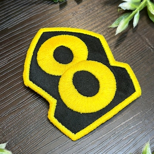 May include: Embroidered patch with a yellow number eight on a black background. The number is outlined in yellow, and the patch has a unique geometric shape. Suitable for clothing or accessories.