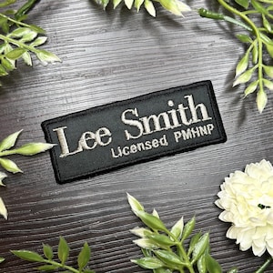 Puede incluir: Parche rectangular negro bordado con el nombre "Lee Smith" en una fuente estilizada. Debajo del nombre, se muestra el texto "Licensed PMHNP". El parche está rodeado de vegetación decorativa y una flor blanca.