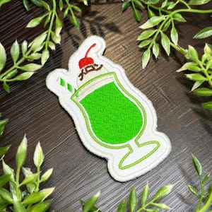 May include: Embroidered patch featuring a green soda drink with whipped cream and a cherry on top. The patch has a white border and a green straw. The Japanese text is visible on the whipped cream.