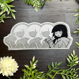 May include: A rectangular patch featuring a black and white anime-style illustration. The design includes a series of figures with light hair, and a central figure with black hair and a surprised expression. The patch is bordered with a light gray thread.