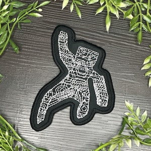May include: A black embroidered patch featuring a white line art design of an astronaut in a space suit. The astronaut is depicted in a dynamic pose with one arm raised. The patch has a thick black border and a detailed, intricate white thread pattern.