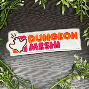 May include: A rectangular embroidered patch with a white background. The text DUNGEON is in orange and MESHI is in pink. To the left, a white ghost-like figure with brown outlines is positioned over a small orange and pink square background.