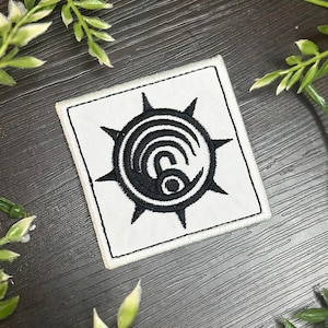 May include: Square white patch with a black embroidered design. The design features a central spiral with a number 6, surrounded by a spiky circular border. The patch has a black border.