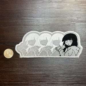 May include: A white embroidered patch featuring a cartoon anime-style character with black hair and a light gray outline. The patch includes three additional faded images of the character. A U.S. quarter is next to the patch for size comparison.