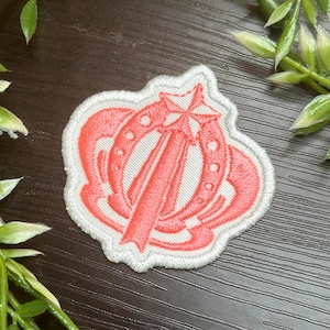 May include: Embroidered patch featuring a coral-colored design of a star, sword, and horseshoe, outlined in white. The patch has a white border and is likely for clothing or accessories.