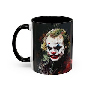 May include: A black and white ceramic mug with a black handle. The mug features a colourful image of the Joker from the film "Joker" with a dark background.