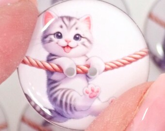 May include: A close-up of a round, clear glass cabochon with a white background and a cartoon image of a gray and white tabby kitten hanging from a rope. The kitten has a pink nose and paws and is smiling.