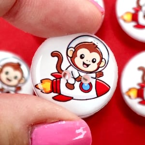 May include: White, round buttons featuring a cartoon monkey astronaut riding a red rocket with orange flames. The monkey wears a spacesuit and helmet. Several buttons are visible against a red background.