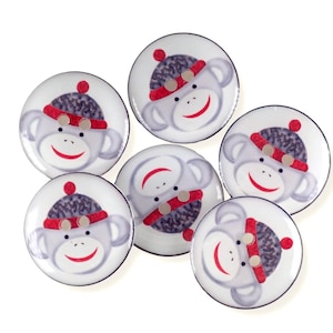 May include: Six white ceramic coasters with a sock monkey design. Each coaster features a grey sock monkey face wearing a grey and red knit hat. The monkey has a red smile and small silver buttons. The coasters are round with a black border.