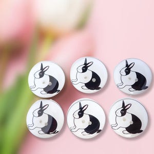 May include: Six white round buttons with black and white rabbit illustrations. The rabbits are sitting with their ears up and their tails behind them.