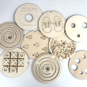 Puede incluir: Una colección de juguetes circulares de madera con varios diseños grabados. Los diseños incluyen una lavadora, zapatos de bebé, dados, flores, una espiral, un laberinto, un tablero de tres en raya y círculos con agujeros. Los juguetes son de madera clara.