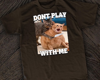 May include: A brown t-shirt with the text "Don't Play With Me" printed in white. The text is above a picture of two cats, one holding a knife to the other's neck.