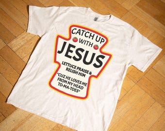 May include: A beige t-shirt with a ketchup-style graphic that says "Catch Up With Jesus." The graphic features two red tomatoes and the text "Lettuce Praise & Relish Him." The text "Cuz He Loves Me From My Head To-Ma-Toes" is also included in the graphic.