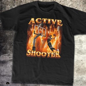 May include: Black t-shirt featuring a graphic of a basketball player performing a slam dunk in a hoop with flames and lightning behind him. The text "ACTIVE SHOOTER" is printed in yellow letters.